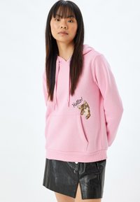 Sudadera rosa con bolsillo frontal, que presenta un diseño de gato y bordado de "¡Hola!". Llevada con una falda de piel sintética negra. Textura suave.