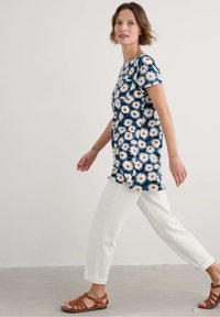 Trumpas rankovės, gėlių rašto tunika su tamsiai mėlynu fonu ir baltomis ramunėmis, kartu su baltomis trumpomis kelnėmis ir rudos spalvos, dirželiais prisiūtomis basutėmis.