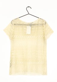 Krämfärgad spetsblus med kort ärm och intrikata geometriska mönster hängande på en svart trådhängare mot en vit bakgrund.