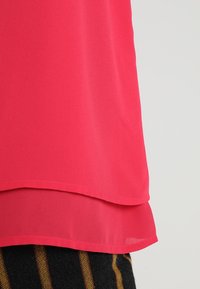 Bright pink layered fabric detail over dark pants with brown vertical stripes, shown against a light gray background.