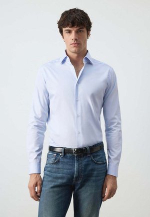 Joven con cabello rizado y oscuro, lleva una camisa azul claro abotonada metida en vaqueros azules con un cinturón negro, de pie frente a un fondo claro.