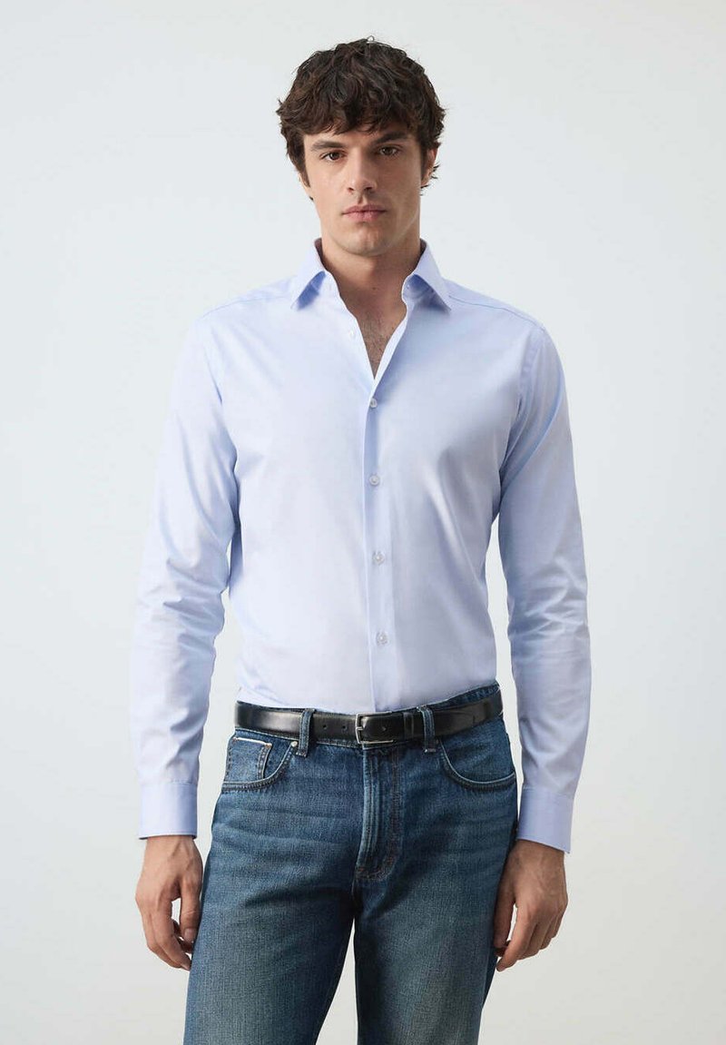 Jeune homme aux cheveux bouclés foncés portant une chemise bleu clair boutonnée, rentrée dans un jean bleu avec une ceinture noire, debout devant un fond clair.