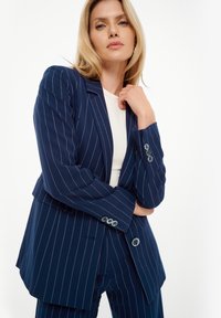 Blazer azul marino a rayas con un ajuste a medida, solapas con muesca, dos botones y una textura de tela suave. Cuenta con un ligero acolchado en los hombros.
