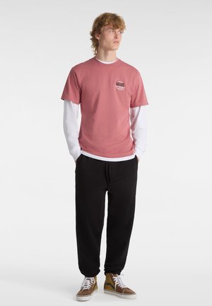 Hellrosa Kurzarm-T-Shirt, über einem weißen Langarmshirt getragen, kombiniert mit schwarzen Jogginghosen und braunen High-Top-Sneakers.