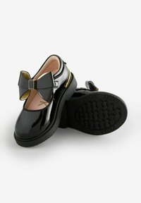 Scarpe in vernice nera con un ampio dettaglio a fiocco, accenti dorati e suole in gomma testurizzata. Design con punta arrotondata e cinturino alla caviglia.