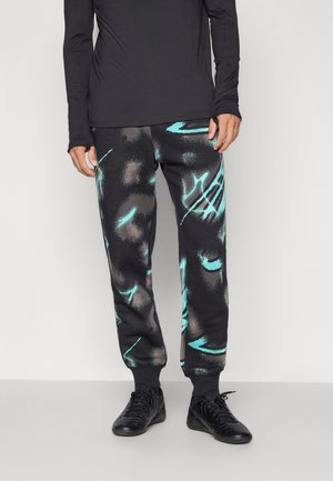 Personne portant un pantalon de jogging noir avec des motifs abstraits turquoise et gris, un t-shirt noir à manches longues, et des chaussures noires à lacets.