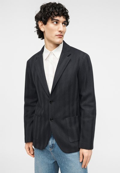 Veste blazer en chevron noire avec deux boutons et poches avant, associée à une chemise blanche et un jean bleu. Coupe ajustée avec un tissu texturé.