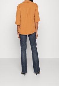 Camisa texturizada de color naranja quemado con mangas de tres cuartos, combinada con jeans de un azul oscuro. La camisa tiene un sutil pliegue en la parte trasera y un dobladillo curvado.