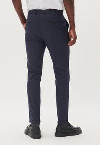 Pantaloni a quadri navy con vestibilità sartoriale, dotati di due tasche posteriori con bottoni e risvolti, indossati con scarpe nere.