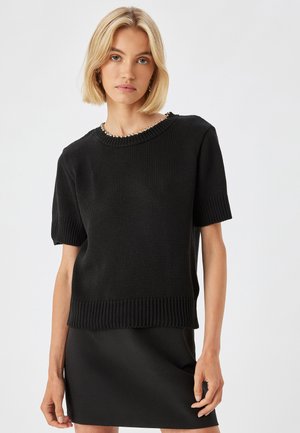 Zwart gebreid sweater met korte mouwen, geribbelde zoom en een brede ronde hals. De textuur is chunky, wat diepte aan het ontwerp toevoegt.