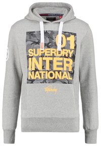 Sudadera con capucha gris claro, con cordones blancos, parche de camuflaje y gran texto amarillo que dice "01 Superdry International" en el frente.