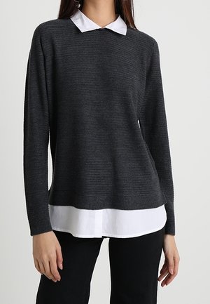 Femme portant un pull en maille côtelée gris foncé superposé à une chemise blanche à col et un pantalon noir, debout devant un fond clair uni.