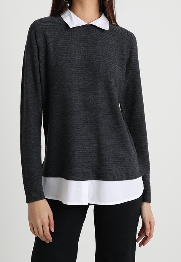 Femme portant un pull en maille côtelée gris foncé superposé à une chemise blanche à col et un pantalon noir, debout devant un fond clair uni.