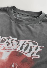 T-shirt gris foncé du groupe Aerosmith avec un texte graphique blanc et rouge délavé et une image du visage d'une personne sous le col.