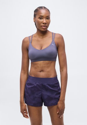 Femme en forme aux cheveux tressés portant un soutien-gorge de sport violet et un short de sport sombre à motif, debout devant un fond clair uni.