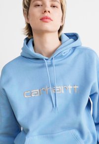 Hanorac albastru deschis dintr-un material moale, cu logo-ul argintiu "carhartt." proeminent pe piept și cu șnururi ajustabile la glugă.