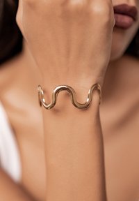 Złota metalowa bransoletka o falistym, falującym wzorze. Gładka tekstura i wypolerowane wykończenie. Noszona na nadgarstku, prezentująca swoją unikalną formę.