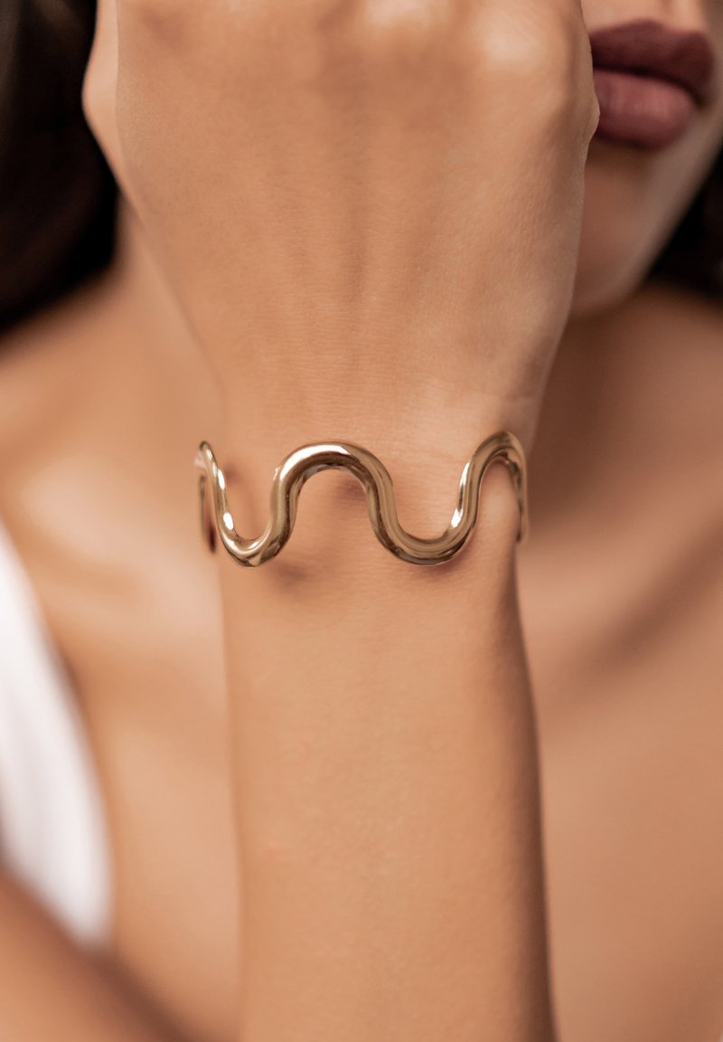 Złota metalowa bransoletka o falistym, falującym wzorze. Gładka tekstura i wypolerowane wykończenie. Noszona na nadgarstku, prezentująca swoją unikalną formę.