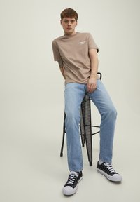 Light brown cotton t-shirt with "Athlete Club" text, paired with light blue straight-leg jeans and black sneakers, sitting on a black metal stool.