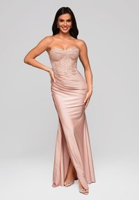 Femme souriante en robe bustier en satin rose poudré avec détails en dentelle, fente haute, et talons scintillants, posant avec une main sur la hanche sur un fond uni.