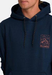 Mężczyzna w granatowej bluzie z kapturem, z czarnymi i żółtymi sznurkami oraz pomarańczowym logo górskim na piersi z napisem "Billabong."