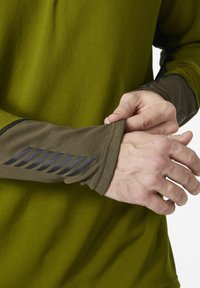 Zelená košile s dlouhým rukávem z texturované látky. Manžety mají tmavě šedou tkaninu s diagonálními pruhy. Detail ruky, jak upravuje rukáv.