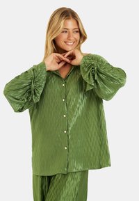 Camisa verde con pliegues, de corte relajado, con botones en la parte delantera y mangas vaporosas. Está hecha de una tela brillante, con una textura de ondas sutil.