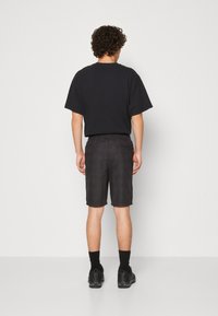 Homme se tenant face à un mur, portant un t-shirt noir ample, un short à carreaux sombre, des chaussettes noires et des chaussures de sport noires.