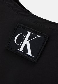 Tessuto nero con una texture liscia presenta un'etichetta quadrata cucita sopra, con la scritta "ck" in carattere serif bianco e un bordo nero.