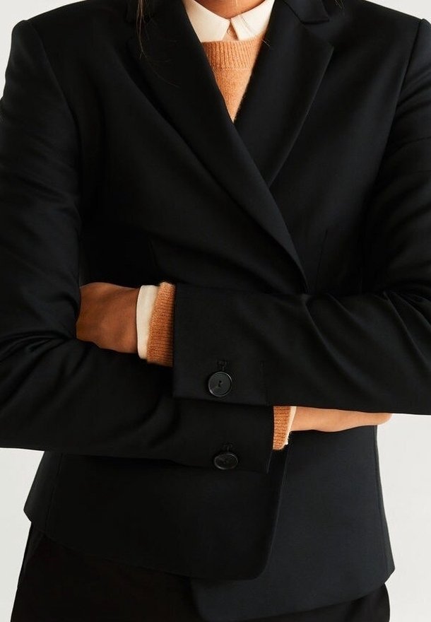 Blazer noir à coupe ajustée, avec deux boutons et manches longues. Un pull orange est visible sous les poignets du blazer.