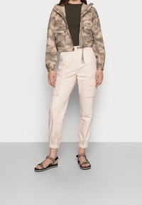 Veste coupée courte et légère avec un motif camouflage marron, associée à un pantalon cargo beige avec des poignets élastiques et des poches latérales, complétée par des sandales à imprimé léopard.