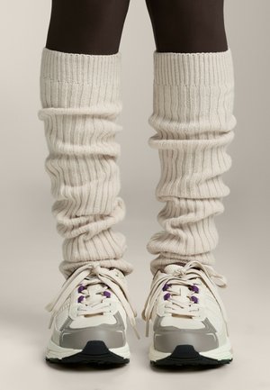 Cremfarbene, gerippte Beinwärmer über dunklen Strumpfhosen, kombiniert mit weißen und grauen Sportschuhen mit lila Akzenten und strukturierten Sohlen.