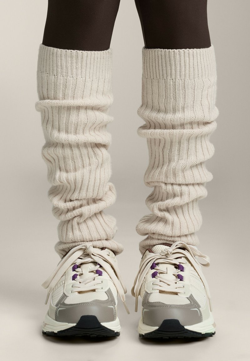 Calze a coste crema indossate sopra collant scuri, abbinate a sneakers sportive bianche e grigie con accenti viola e suole testurizzate.