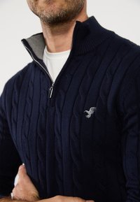 Marine gebreide ritspull met een kabelpatroon en een grijze kraagvoering. Heeft een klein geborduurd logo op de borst. Zachte textuur.