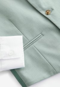 Helles grünes Blazerstoff mit einer glatten Textur, ausgestattet mit einer Brusttasche und einem weißen, strukturierten Hemdmanschette mit einem runden Knopf.
