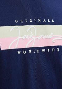 Tmavě modré tričko s horizontálními pruhy v krémové a růžové barvě s bílým vyšitým textem: "Jack Jones" a "ORIGINALS WORLDWIDE."