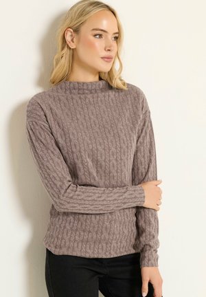 Blond kvinde iført en mauve kabelstrik langærmet trøje og sorte bukser, stående op ad en lys ensfarvet væg, og kigger sidelæns.