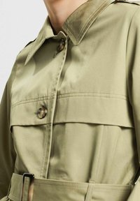 Jasnooliwkowa kurtka z gładkiego materiału, posiadająca kołnierzyk, dużych guzik oraz pasek w talii z wyraźnie zdefiniowaną konstrukcją.