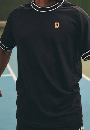 Person iført sort sportstrøje med hvid kant og lille rød og gul logo på brystet står på en blå tennisbane.