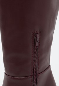 Botte en cuir bordeaux hautes jusqu'au genou avec une finition lisse, dotée d'une fermeture zippée sur le côté et d'un contraste de couture le long des coutures.
