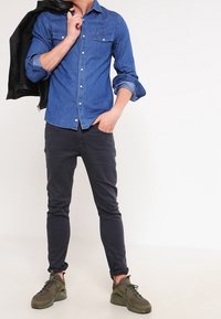 Sininen farkkupaita, jossa on kaksi rintataskua, yhdistetty tummiin kapea-mitoitettuihin housuihin ja oliivinvihreisiin lenkkareihin. Henkilö pitää mustaa takkia.