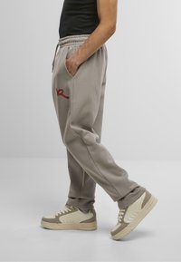 Graue Jogginghosen mit elastischem Bund, seitlichen Taschen und einem roten gestickten Logo. Kombiniert mit beigefarbenen und cremefarbenen Sneakers mit strukturierten Akzenten.