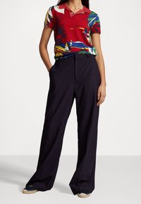 Kvinna klädd i en röd polotröja med tropiskt mönster, svarta vida byxor och svarta espadriller med beige sula, stående med händerna i fickorna.