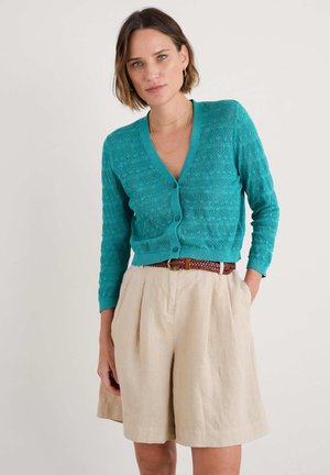 Teal neulecardigan, jossa on V-pääntie, edessä napit ja tekstuuri-kuvioita; yhdistetty vaaleanbeigeihin korkeavyötäröisiin shortseihin ja punottuun vyöhön.