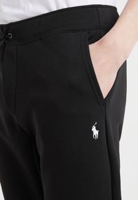 Svarta sweatpants med en slät textur, med framfickor och en vit polo-logotyp på vänster lår. Elastisk midja med snörning.
