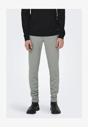 Mann trägt schlanke, hellgraue Hosen, ein schwarzes Langarmoberteil und schwarze Lederstiefel, steht vor einem schlichten, hellen Hintergrund.