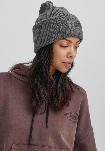 Pelēks adīts cepure ar ribota tekstūru un apmalīti, ar tumšu logo uzlīmi. Vilkts kopā ar brūnu kapuci, uz kura redzams sānu logo.