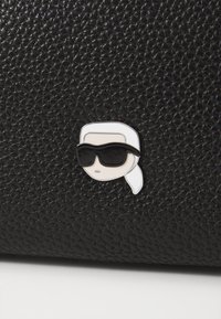 Sac en cuir noir texturé avec une épingle en argent représentant une figure portant des lunettes de soleil et ayant des cheveux blancs. Les accents incluent une finition métallique lisse.