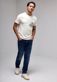 Camiseta crema con bolsillo que presenta un diseño gráfico abstracto en negro, combinada con jeans azul oscuro y zapatillas blancas con suelas marrones.