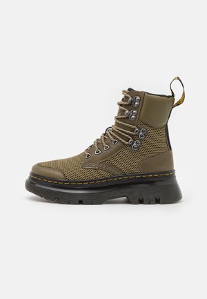 Olijfgroen mesh veterschoen met zwarte chunky zool, metalen oogjes, gele stiksels en een treklus bij de enkel op een witte achtergrond.